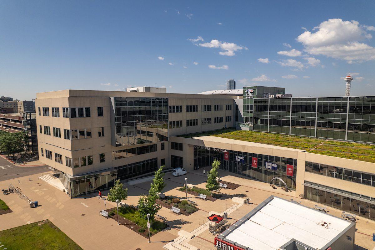 The Jordan Student Success Building on the MSU Denver campus, featuring a modern design with large glass windows and a green roof. The building surrounds a courtyard with trees, benches, and pathways. The Denver skyline and a tower are visible in the background under a clear blue sky.